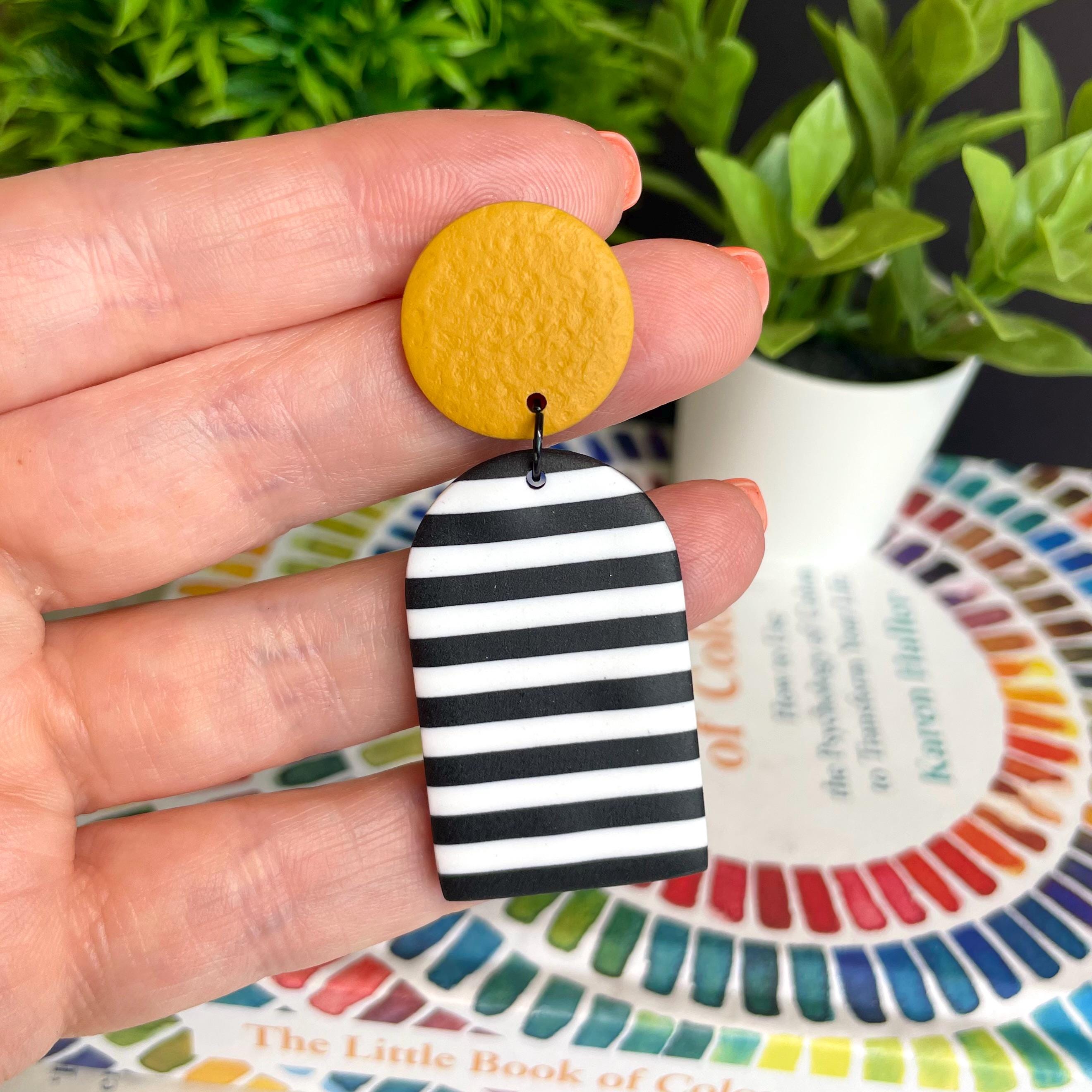Hand holding a yellow and black striped earring for size reference with a plant and colourful book in the background