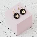 A pair of gold hoop earrings with a black polymer clay disc and a small gold heart, displayed on a light pink cube against a white speckled background.
