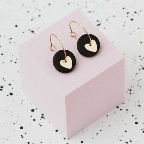 A pair of gold hoop earrings with a black polymer clay disc and a small gold heart, displayed on a light pink cube against a white speckled background.