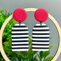 Pair of earrings with red circular tops and black and white striped bottoms, displayed against a green plant background.