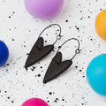 A pair of handmade black heart-shaped hoop earrings on black hoops, displayed on a white speckled surface surrounded by colourful polymer clay balls.
