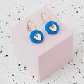 A pair of gold hoop earrings with a blue polymer clay disc and a small gold heart, displayed on a light pink cube against a white speckled background.