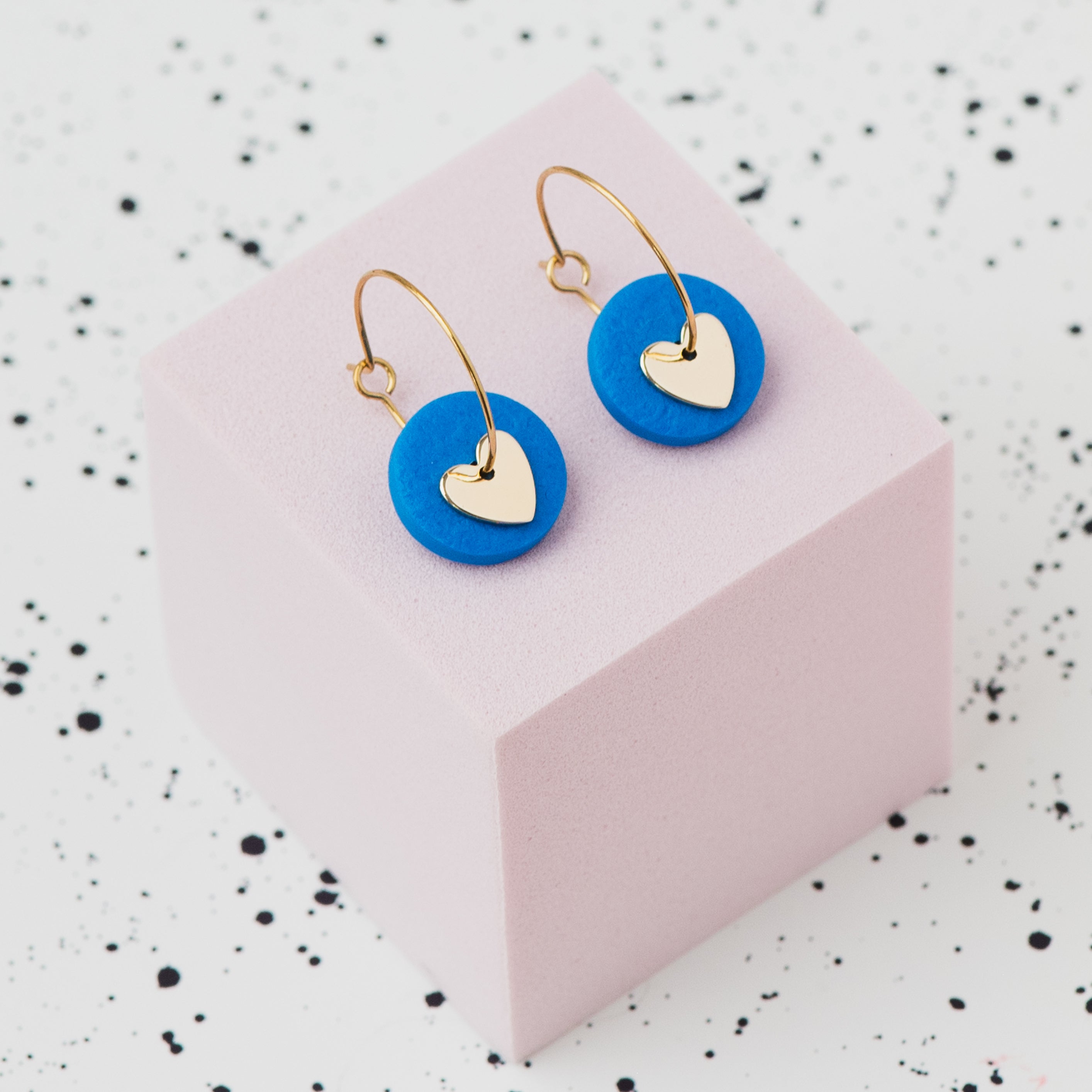A pair of gold hoop earrings with a blue polymer clay disc and a small gold heart, displayed on a light pink cube against a white speckled background.