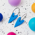 A pair of handmade blue heart-shaped hoop earrings on black hoops, displayed on a white speckled surface surrounded by colourful polymer clay balls.