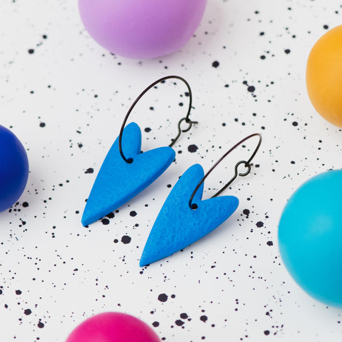 A pair of handmade blue heart-shaped hoop earrings on black hoops, displayed on a white speckled surface surrounded by colourful polymer clay balls.