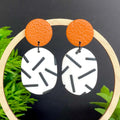 Earrings with burnt orange top and mono sprinkle oval bottom , set against a dark background with green foliage.