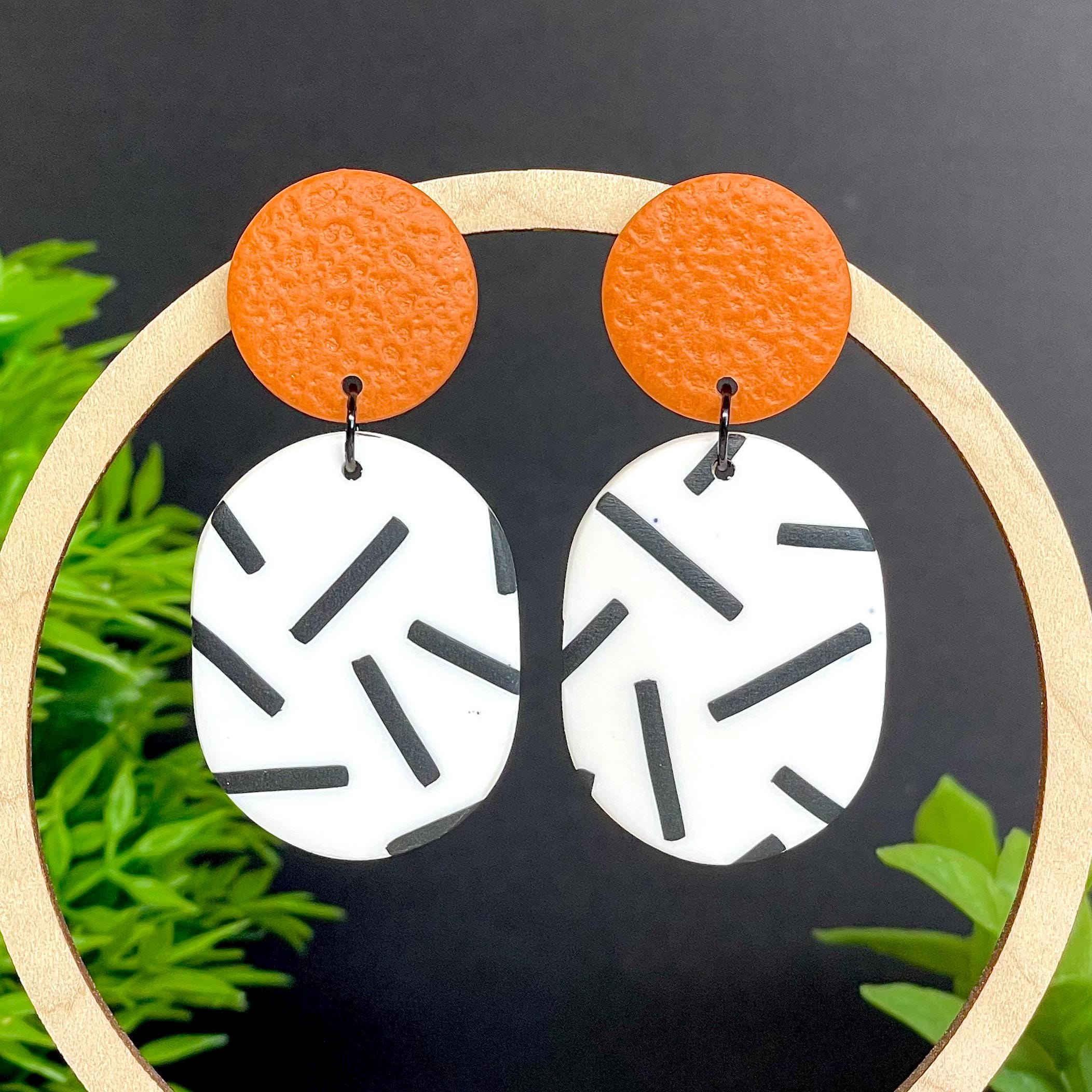 Earrings with burnt orange top and mono sprinkle oval bottom , set against a dark background with green foliage.