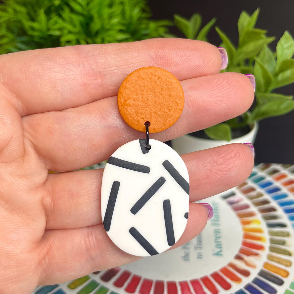 Hand holding the burnt orange top and mono sprinkle oval bottom earrings , with a plant and colour wheel in the background.