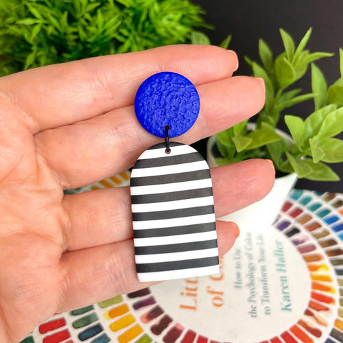 Hand holding a blue and black and white striped earring with plants in the background