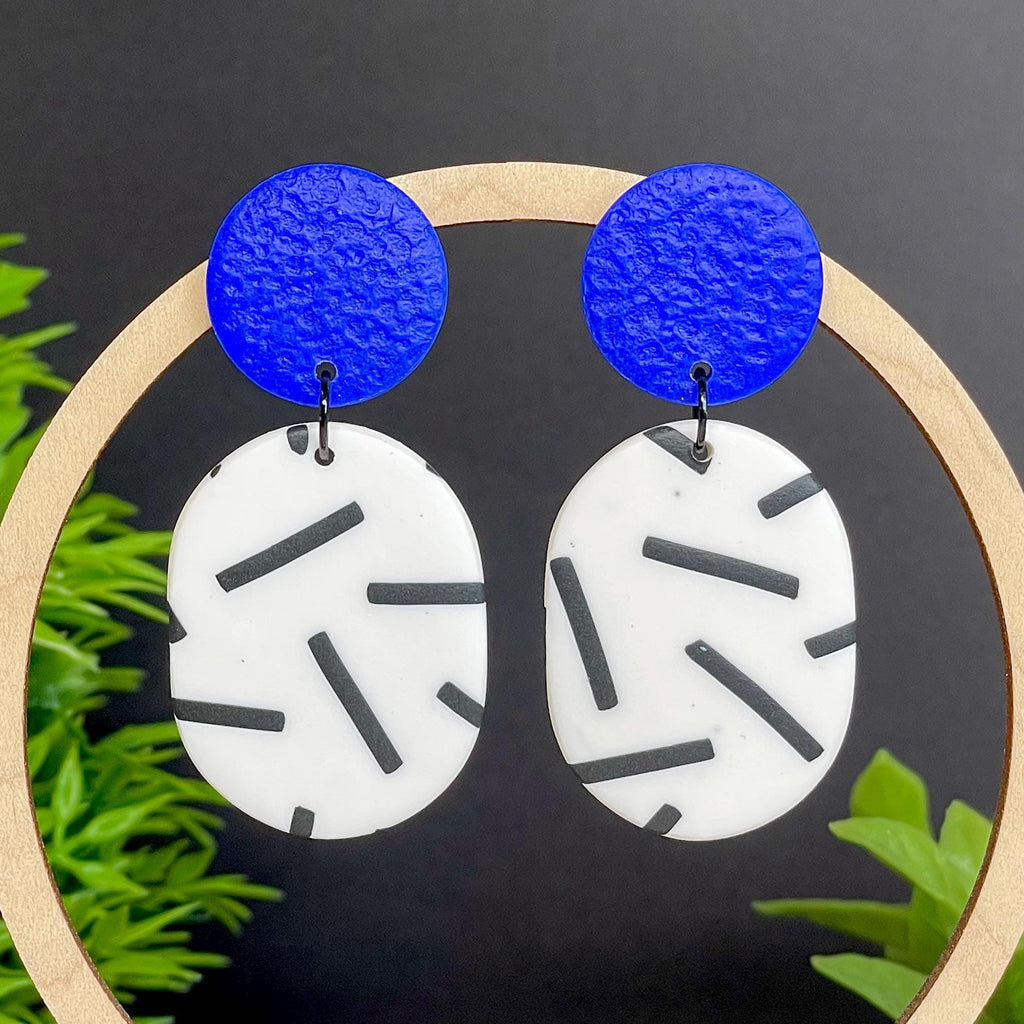 Earrings with electric blue circular tops and monochrome sprinkles oval bottoms, set against a dark background with green leaves.
