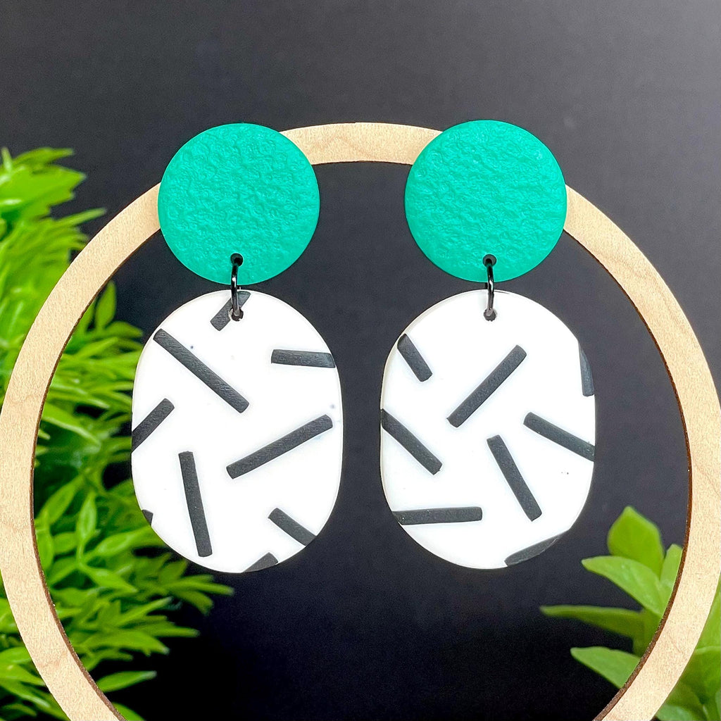 Earrings with emerald green tops & monochrome sprinkle oval bottoms, displayed on a wooden hoop against a dark background with greenery.