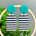Black and white striped earrings with green circular tops on a wooden stand against a green leafy background.
