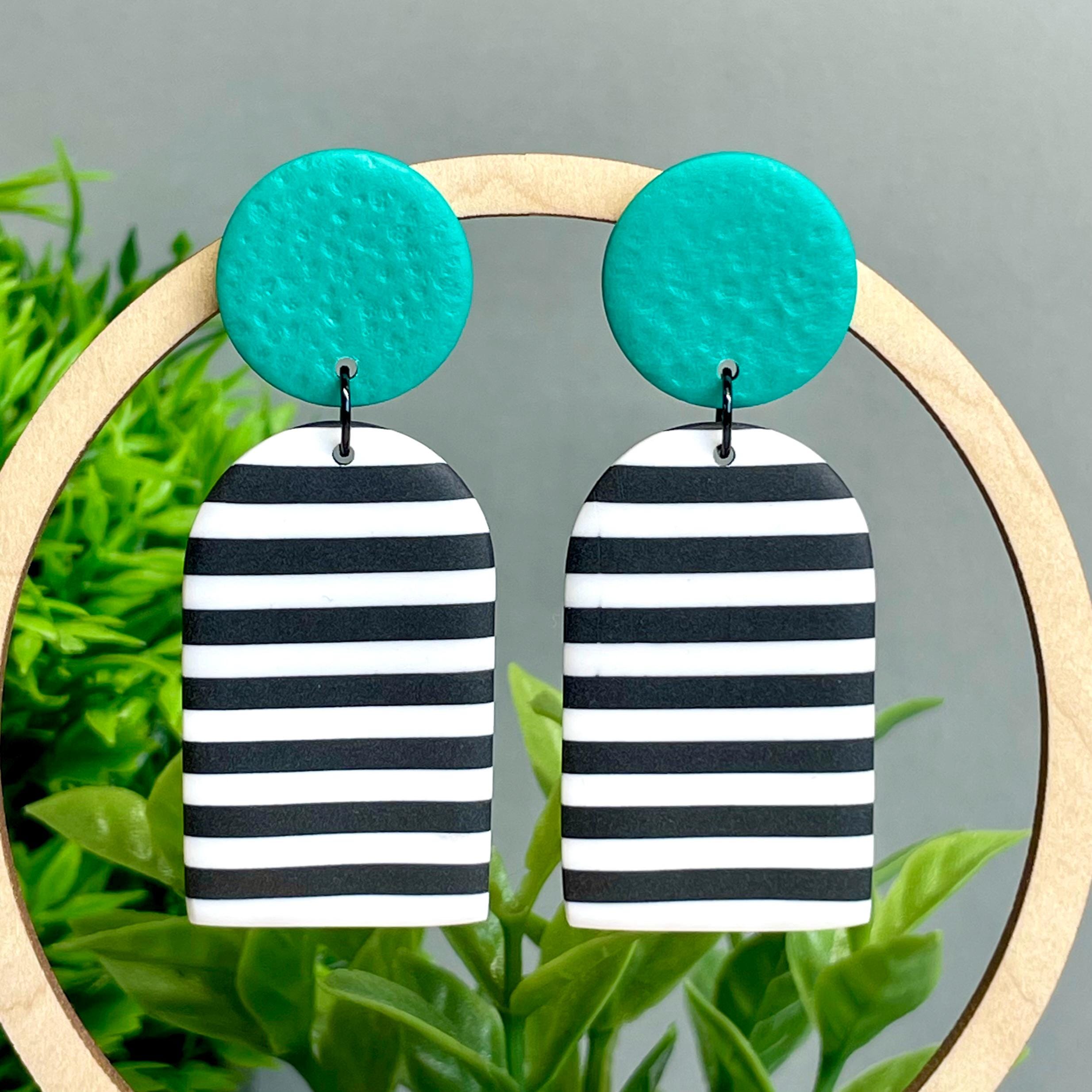 Black and white striped earrings with green circular tops on a wooden stand against a green leafy background.