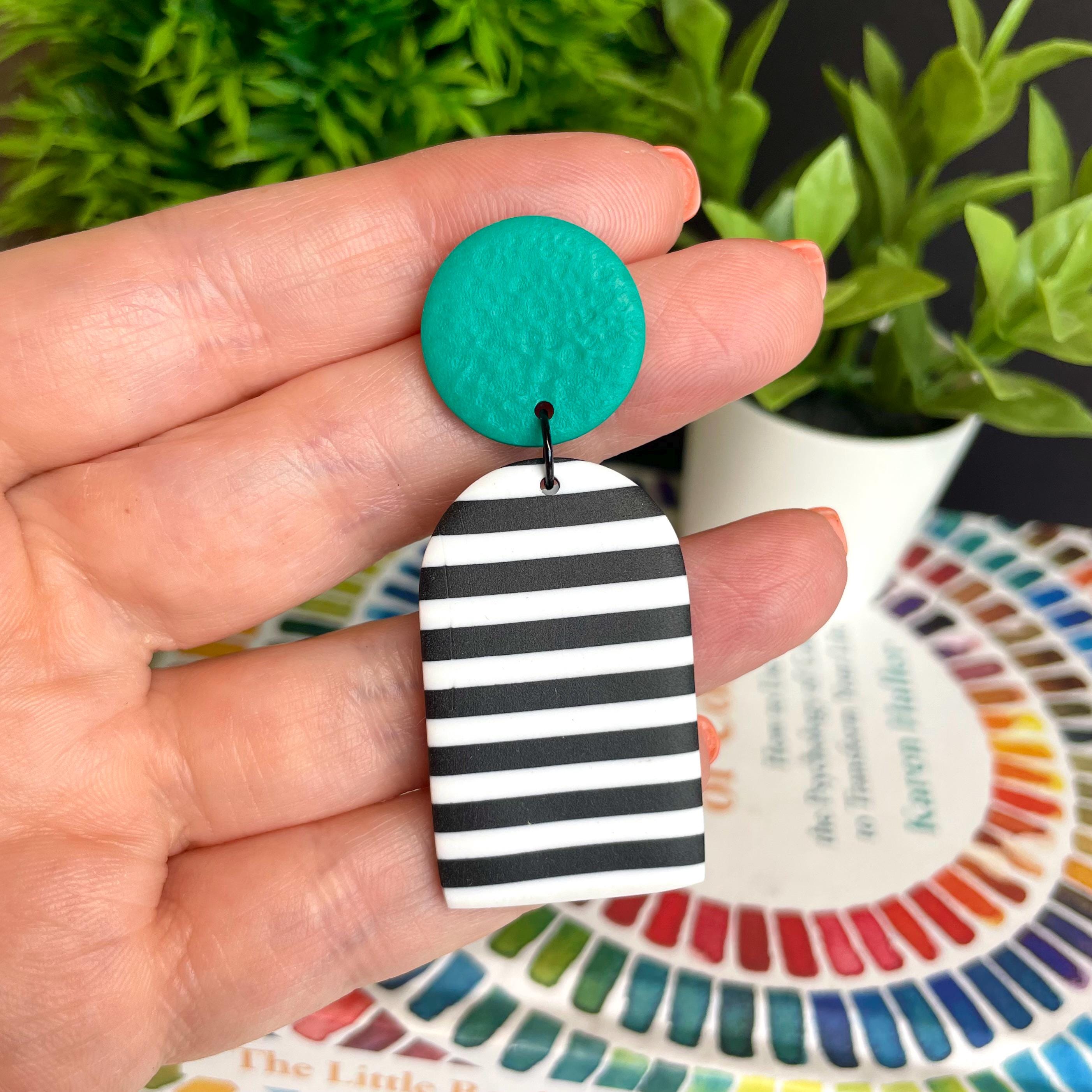 Hand holding a green and black and white striped earring with a plant and color wheel in the background.
