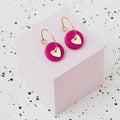 A pair of gold hoop earrings with a metallic bronze polymer clay disc and a small gold heart, displayed on a light pink cube against a white speckled background.