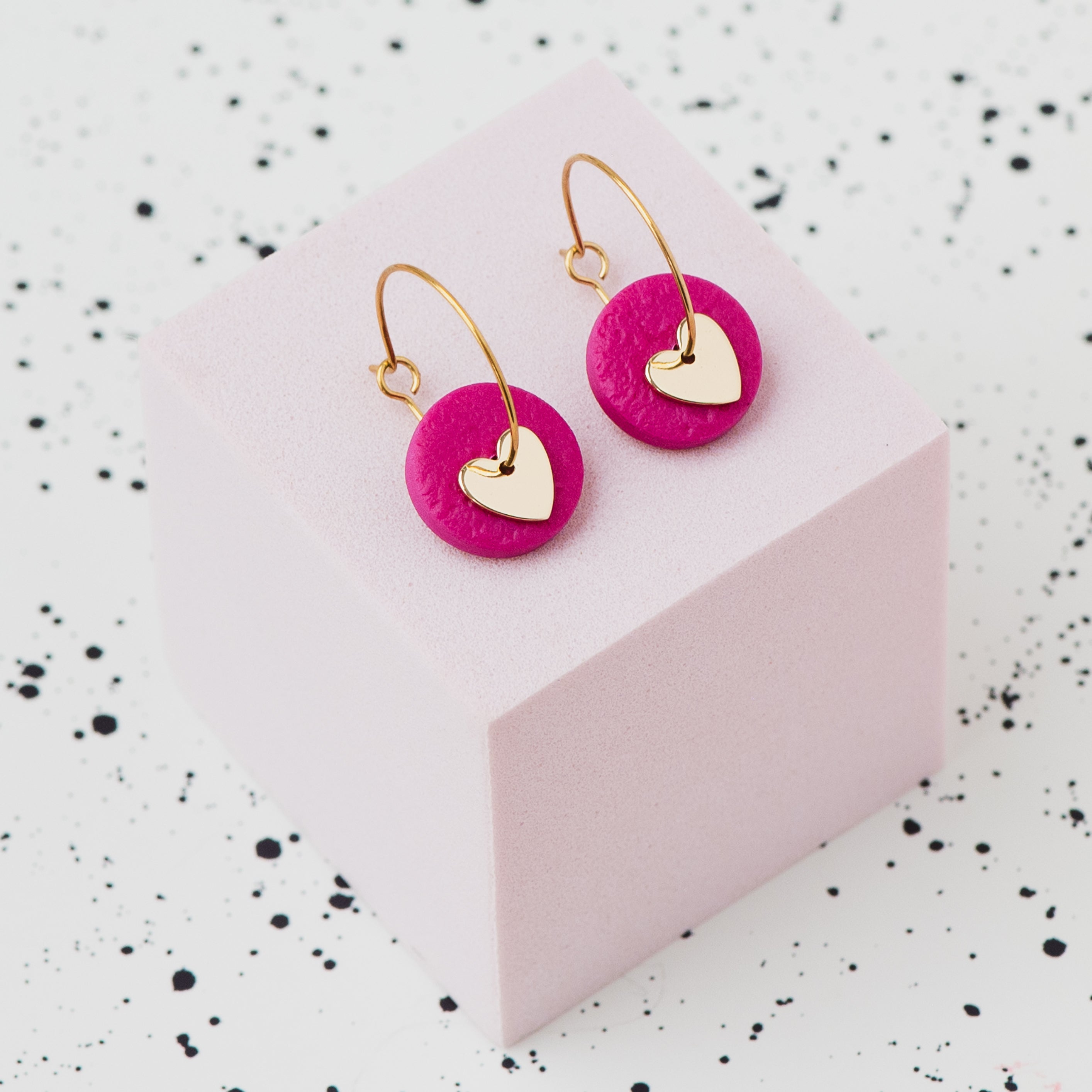 A pair of gold hoop earrings with a metallic bronze polymer clay disc and a small gold heart, displayed on a light pink cube against a white speckled background.