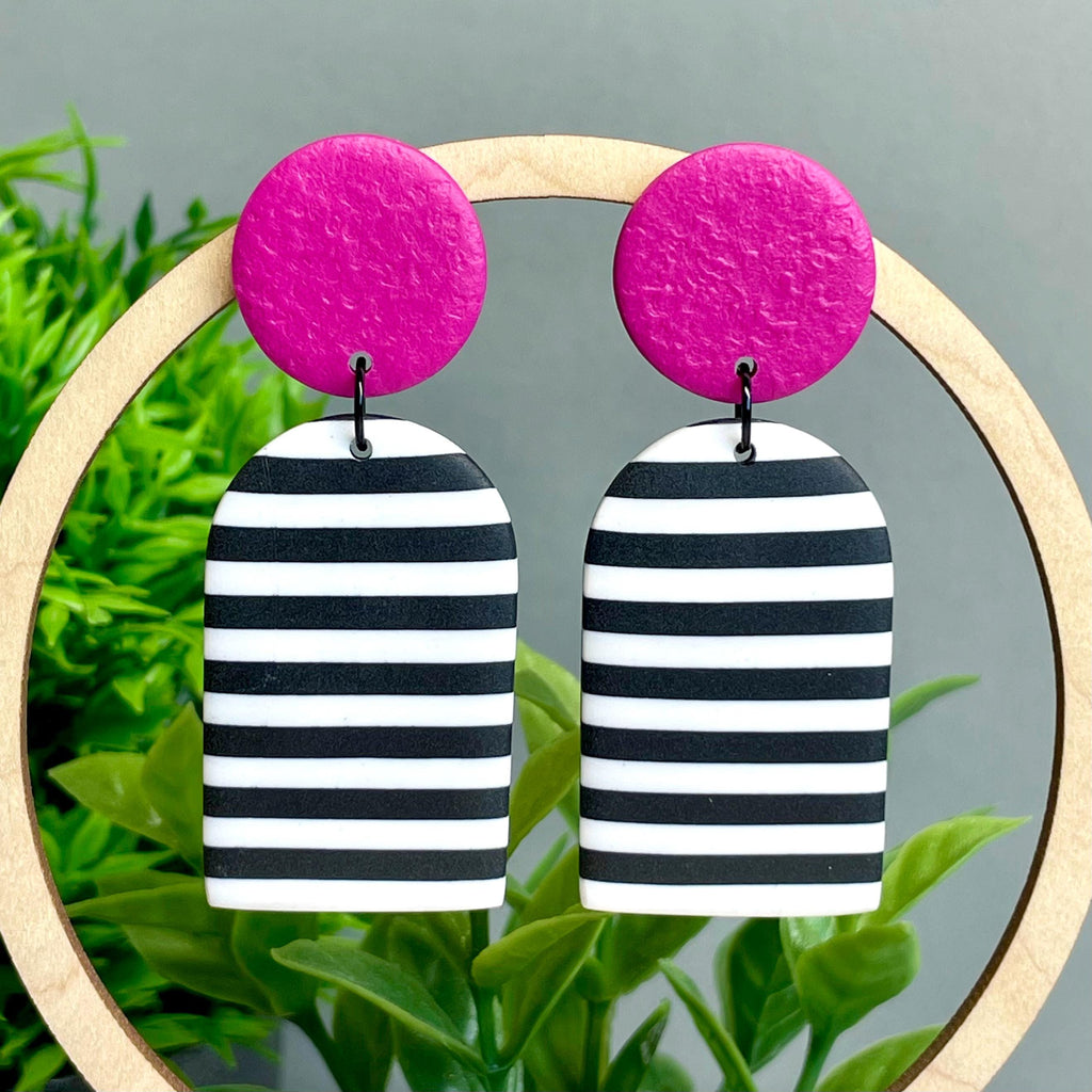 Pair of earrings with pink top and black and white striped bottom hanging on a wooden hoop against a green plant background.