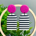 Pair of earrings with pink top and black and white striped bottom hanging on a wooden hoop against a green plant background.