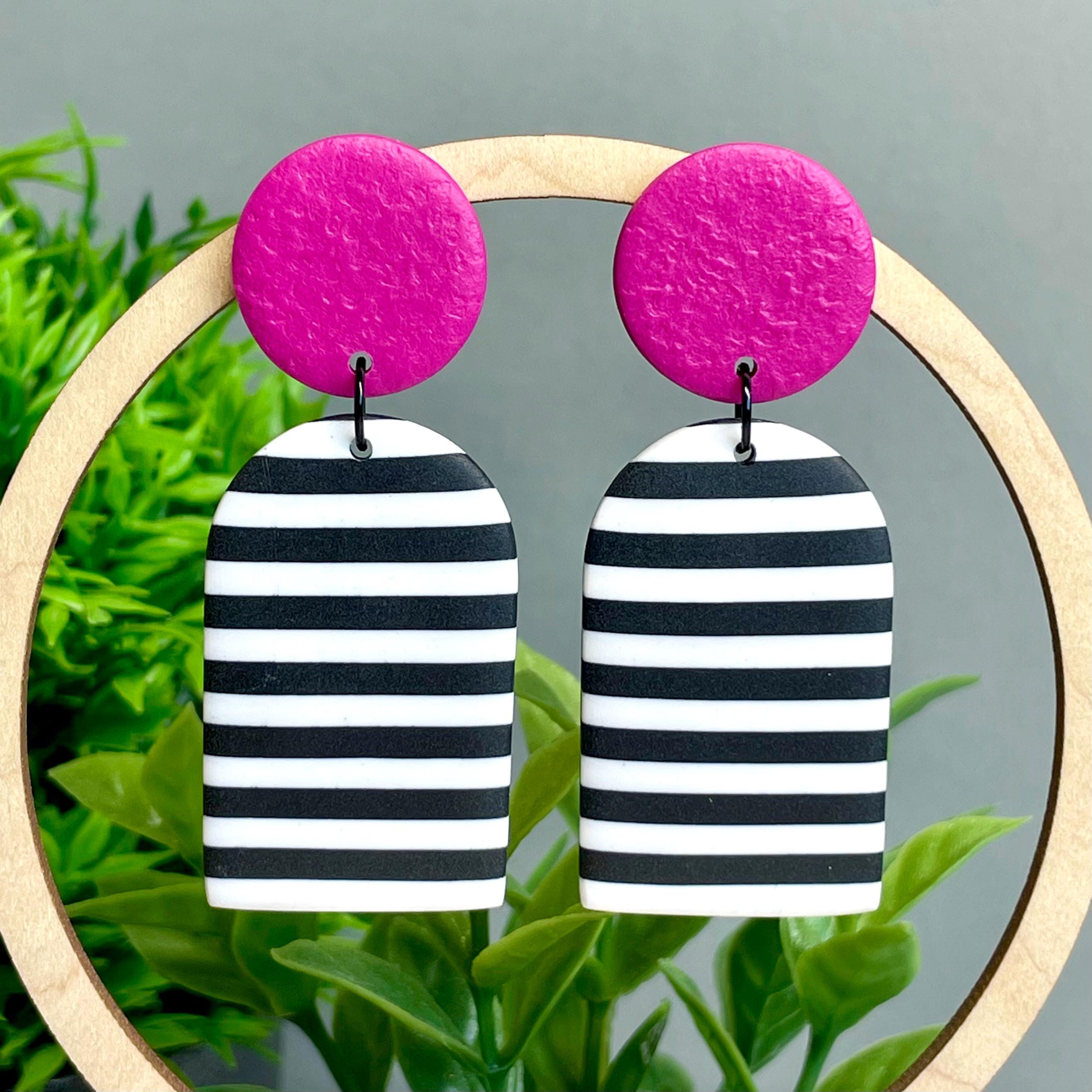 Pair of earrings with pink top and black and white striped bottom hanging on a wooden hoop against a green plant background.