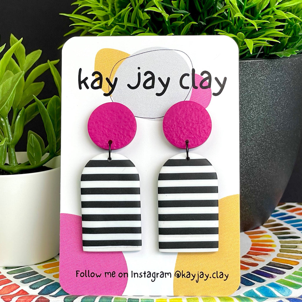 Pair of black & white striped earrings with pink top on a 'kay jay clay' branded card with plants in the background.