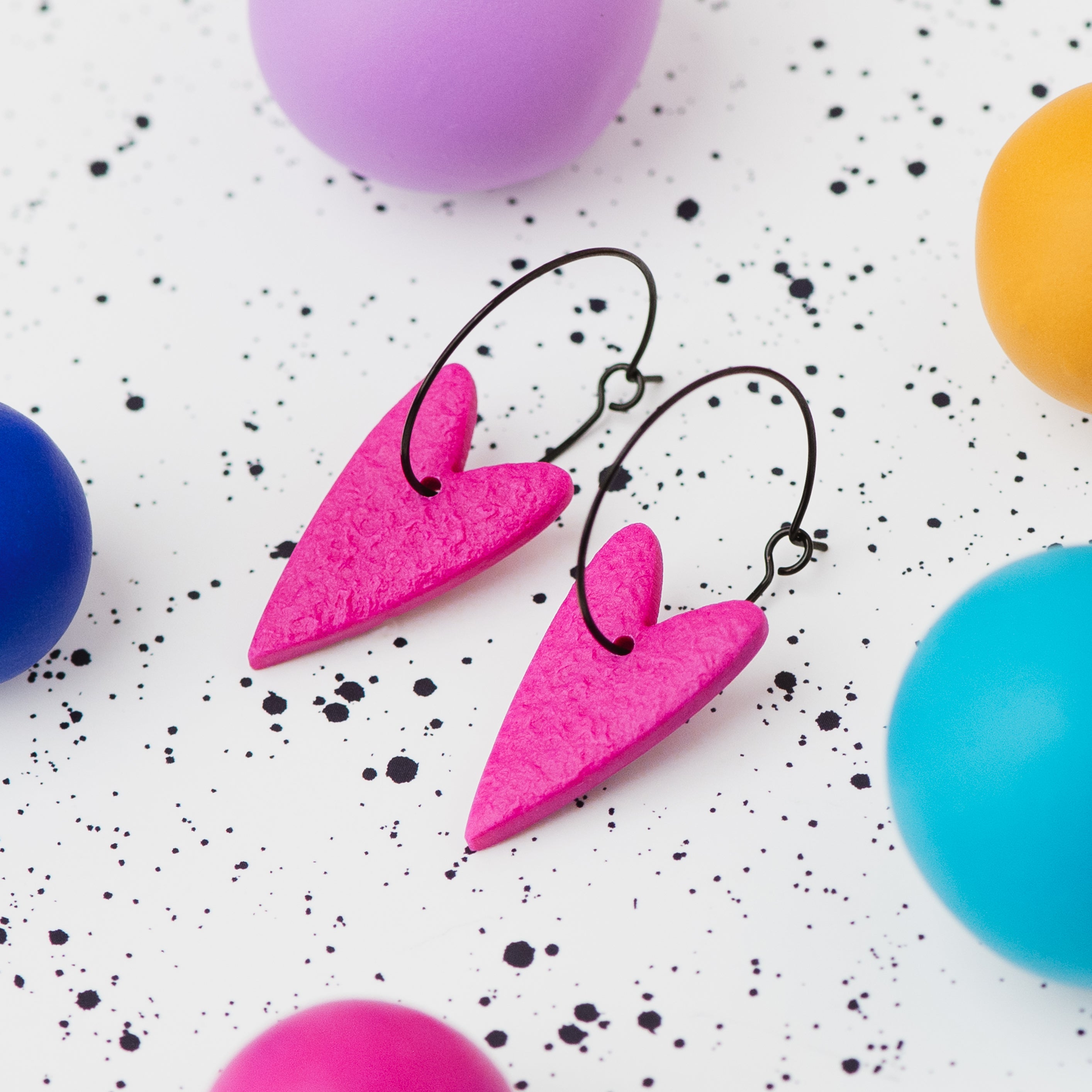 A pair of handmade hot pink heart-shaped hoop earrings on black hoops, displayed on a white speckled surface surrounded by colourful polymer clay balls.