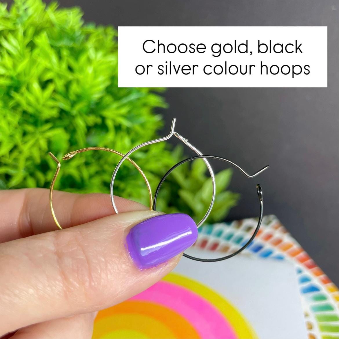 Three hoop earrings in gold, silver, and black held by a hand to show colour options available