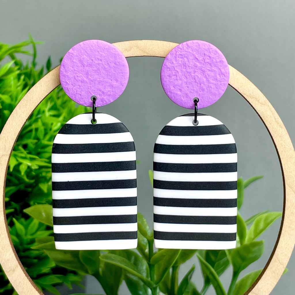 Black and white striped earrings with lilac purple circular tops on a green plant background