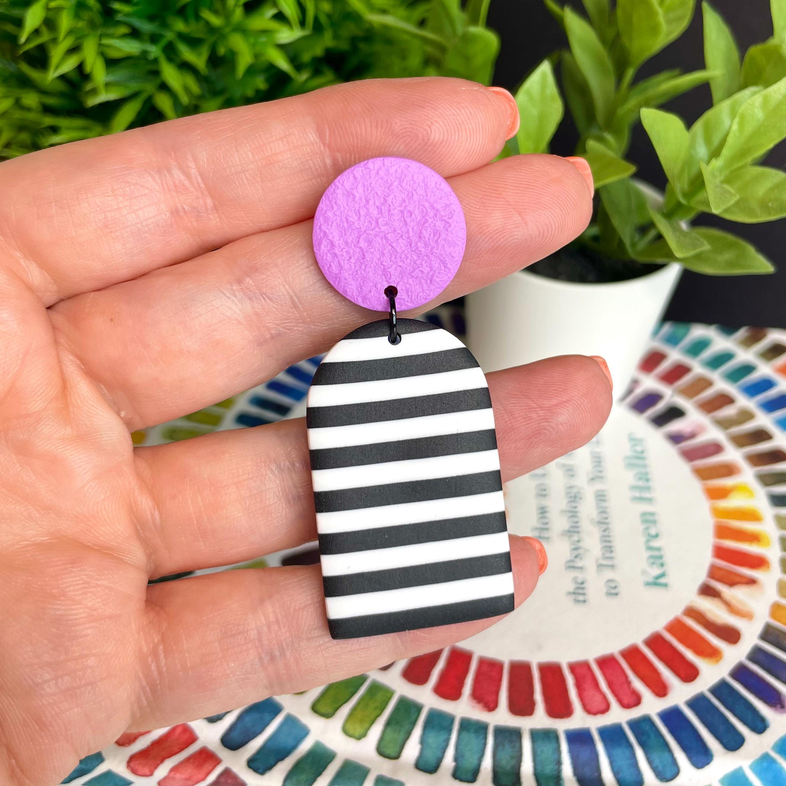 Hand holding a black and white striped earring with a purple circle, with a color swatch book in the background.