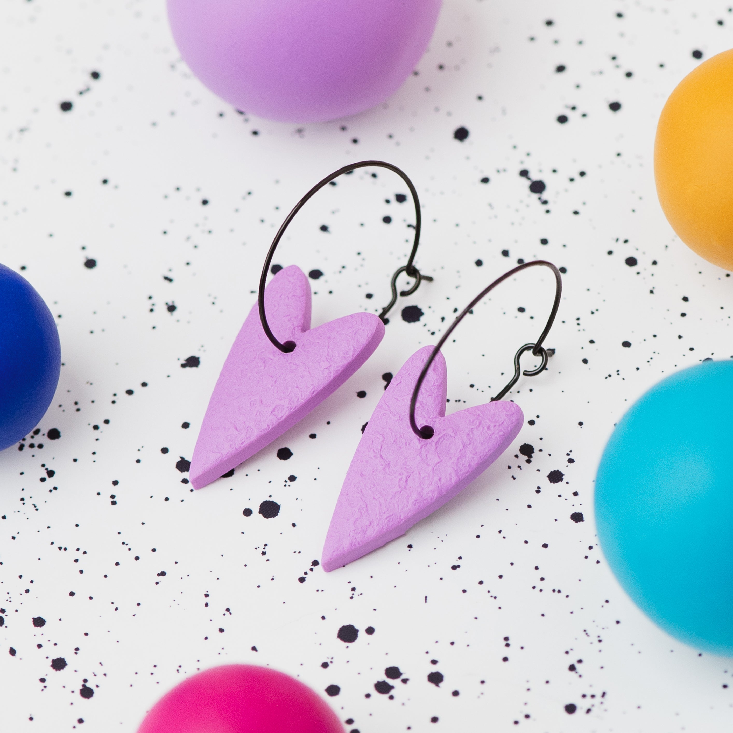 A pair of handmade lilac heart-shaped hoop earrings on black hoops, displayed on a white speckled surface surrounded by colourful polymer clay balls.