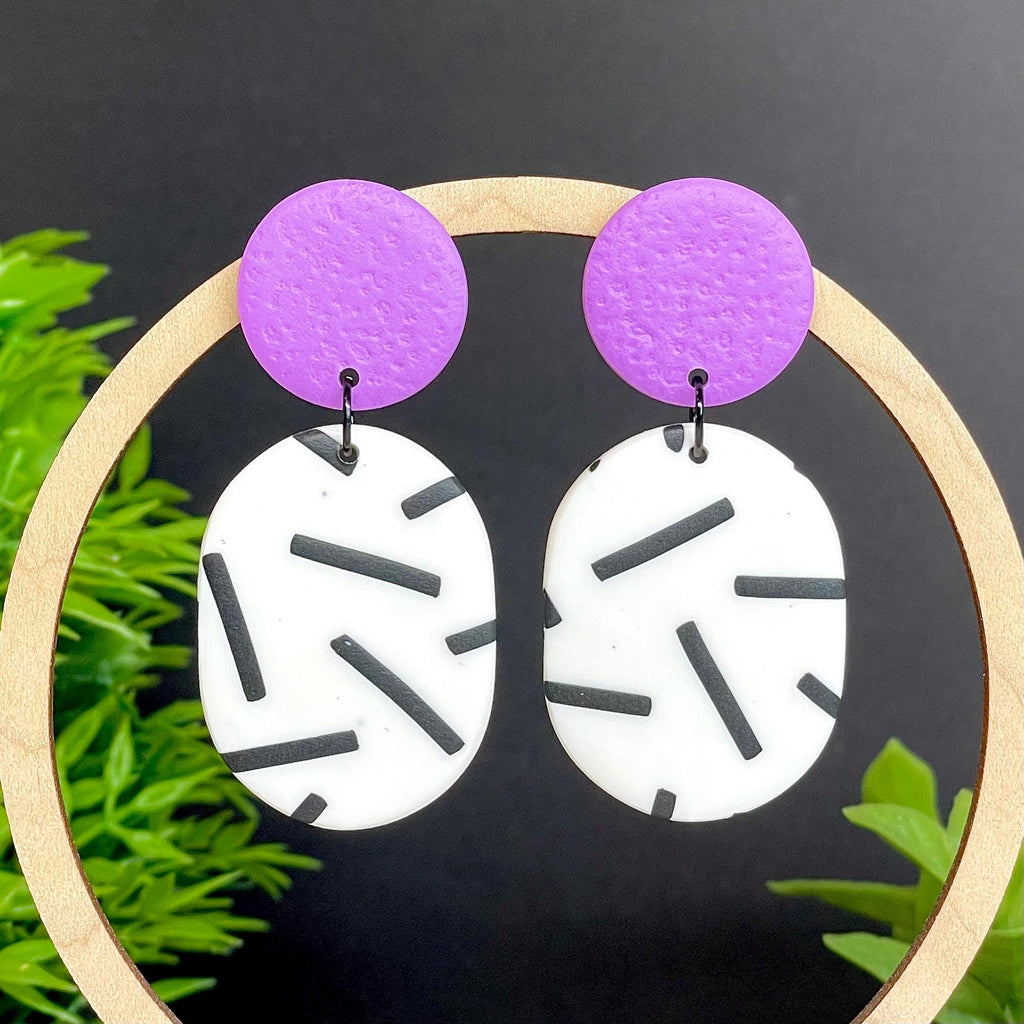 Earrings with lilac tops & monochrome sprinkle oval bottoms, displayed against a dark background with greenery.