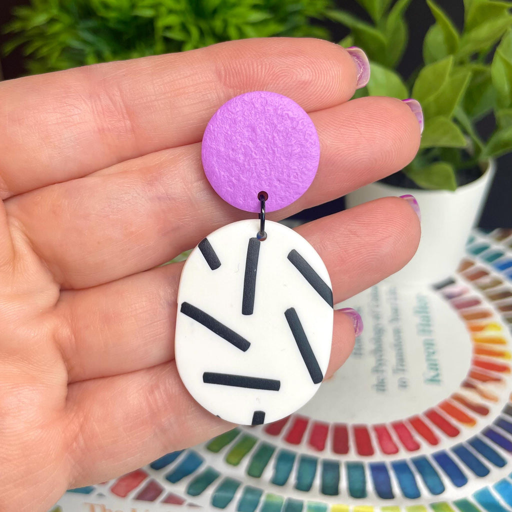 Hand holding a pair of earrings with a lilac top & monochrome sprinkle oval bottoms, against a colourful background.