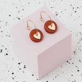 A pair of gold hoop earrings with a metallic bronze polymer clay disc and a small gold heart, displayed on a light pink cube against a white speckled background.