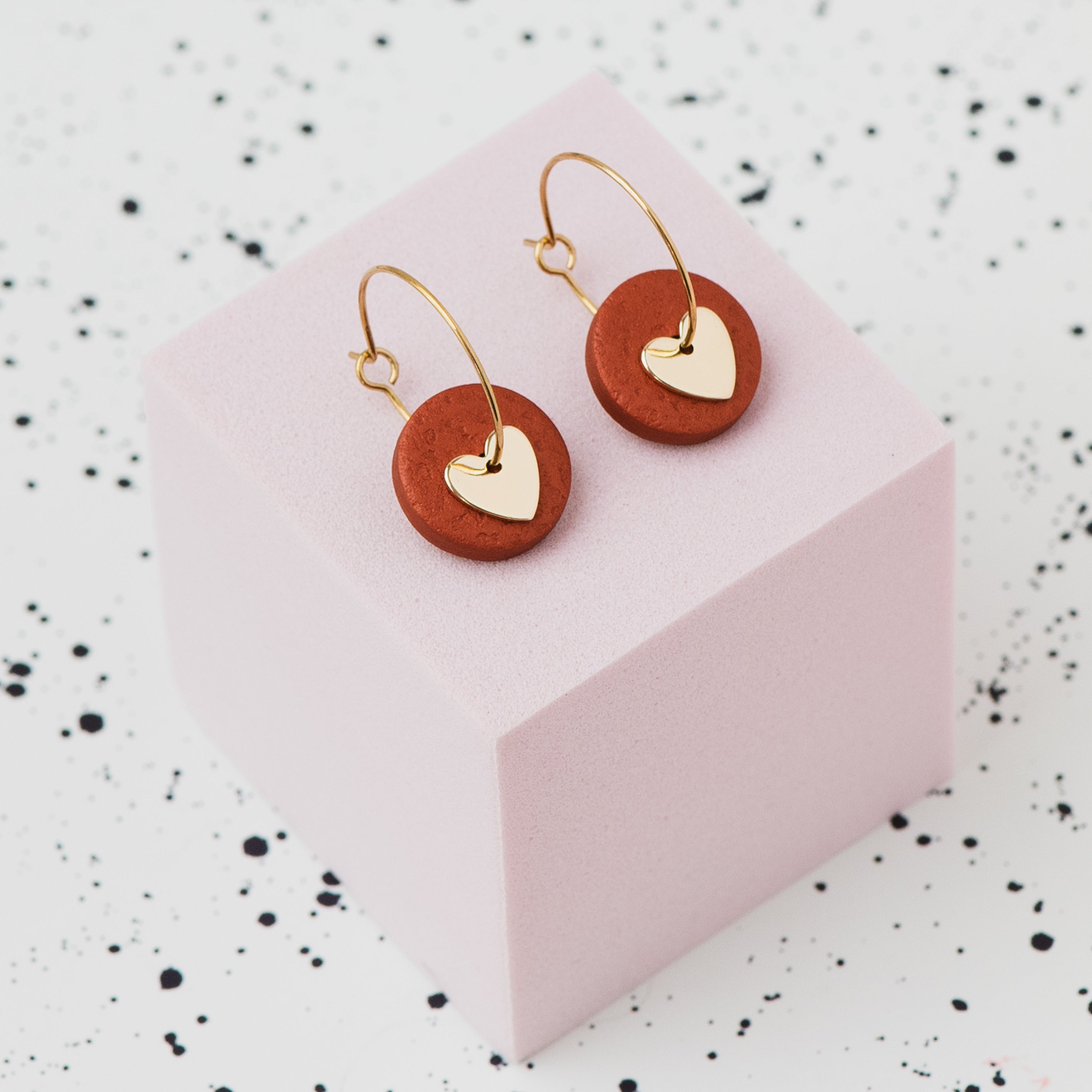 A pair of gold hoop earrings with a metallic bronze polymer clay disc and a small gold heart, displayed on a light pink cube against a white speckled background.