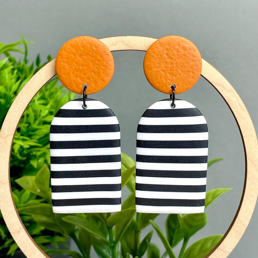 Black and white striped earrings with orange circular tops on a wooden hoop against a green plant background.