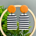 Black and white striped earrings with orange circular tops on a wooden hoop against a green plant background.