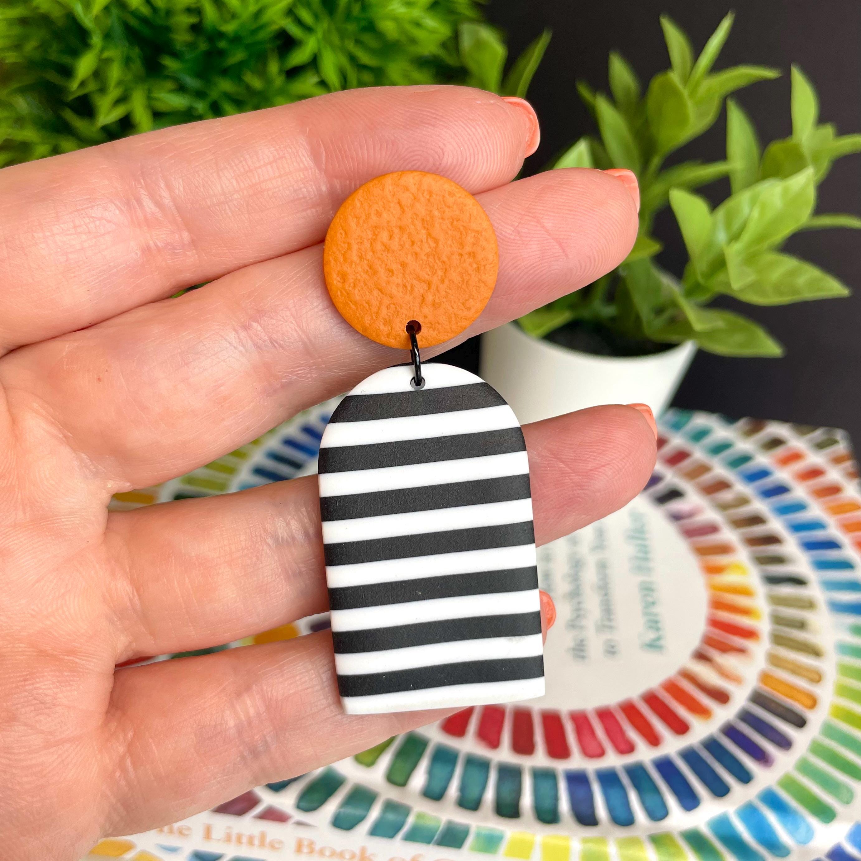 Hand holding a pair of earrings with an orange top and black and white striped bottom, with a colorful book and plant in the background.