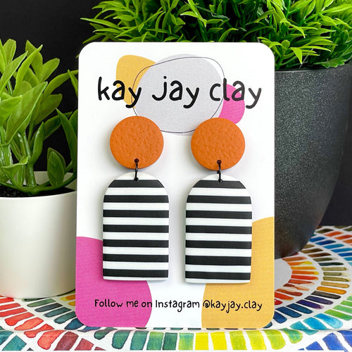 Monochrome stripes dangle earrings with orange top on a 'kay jay clay' branded card with plants in the background
