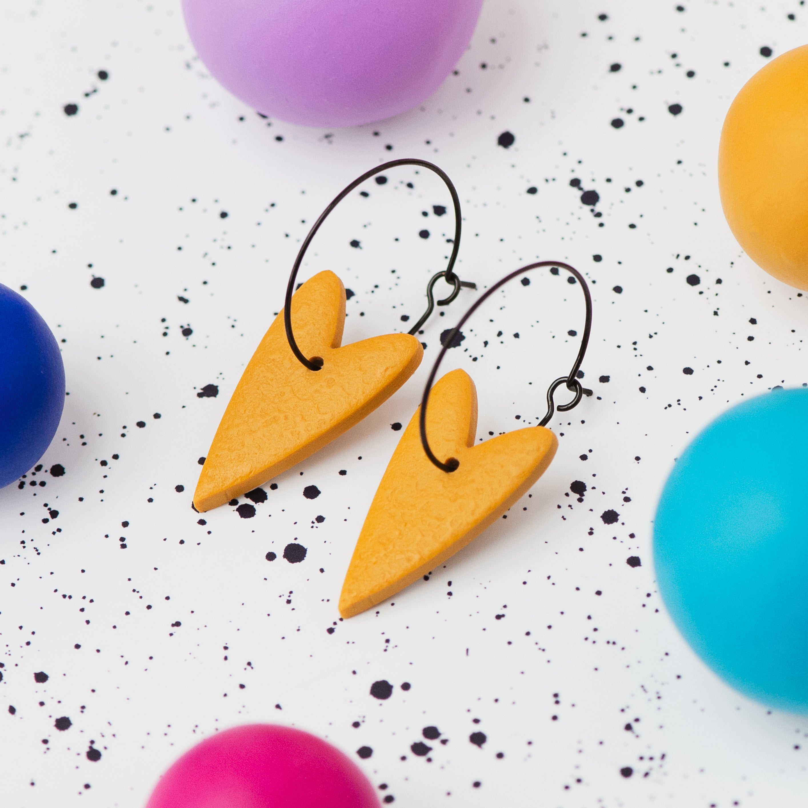 A pair of handmade mustard yellow heart-shaped hoop earrings on black hoops, displayed on a white speckled surface surrounded by colourful polymer clay balls.