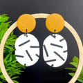 Pair of earrings with mustard yellow top & monochrome sprinkle oval bottoms, displayed on a wooden hoop against a dark background with green leaves.