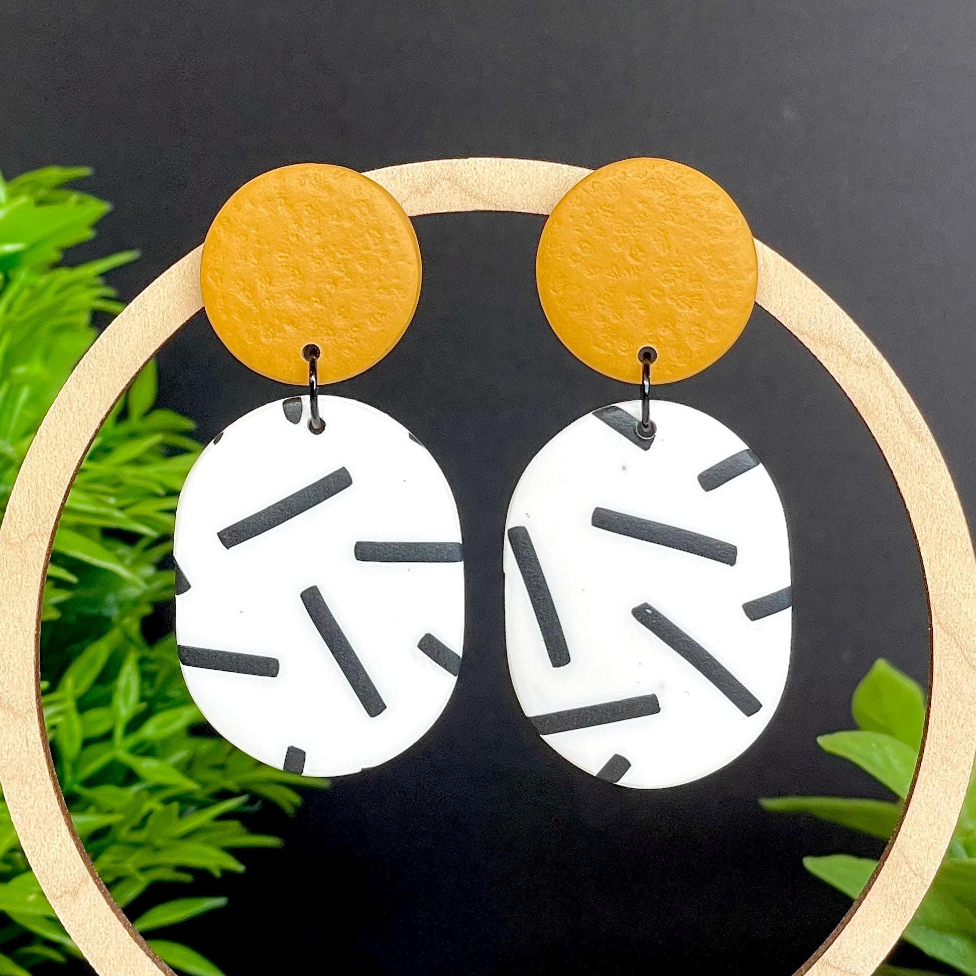 Pair of earrings with mustard yellow top & monochrome sprinkle oval bottoms, displayed on a wooden hoop against a dark background with green leaves.