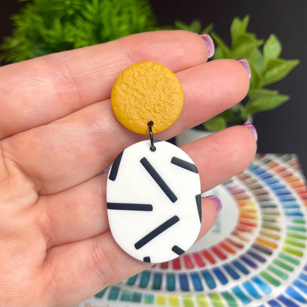Hand holding an earring with mustard yellow top & monochrome sprinkle oval bottom against a colourful background