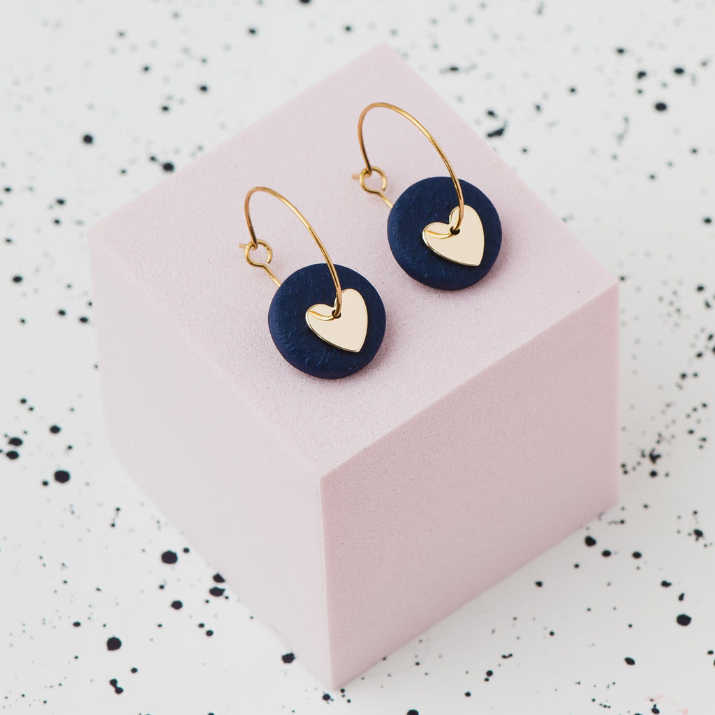 A pair of gold hoop earrings with a navy blue polymer clay disc and a small gold heart, displayed on a light pink cube against a white speckled background.
