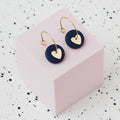 A pair of gold hoop earrings with a navy blue polymer clay disc and a small gold heart, displayed on a light pink cube against a white speckled background.