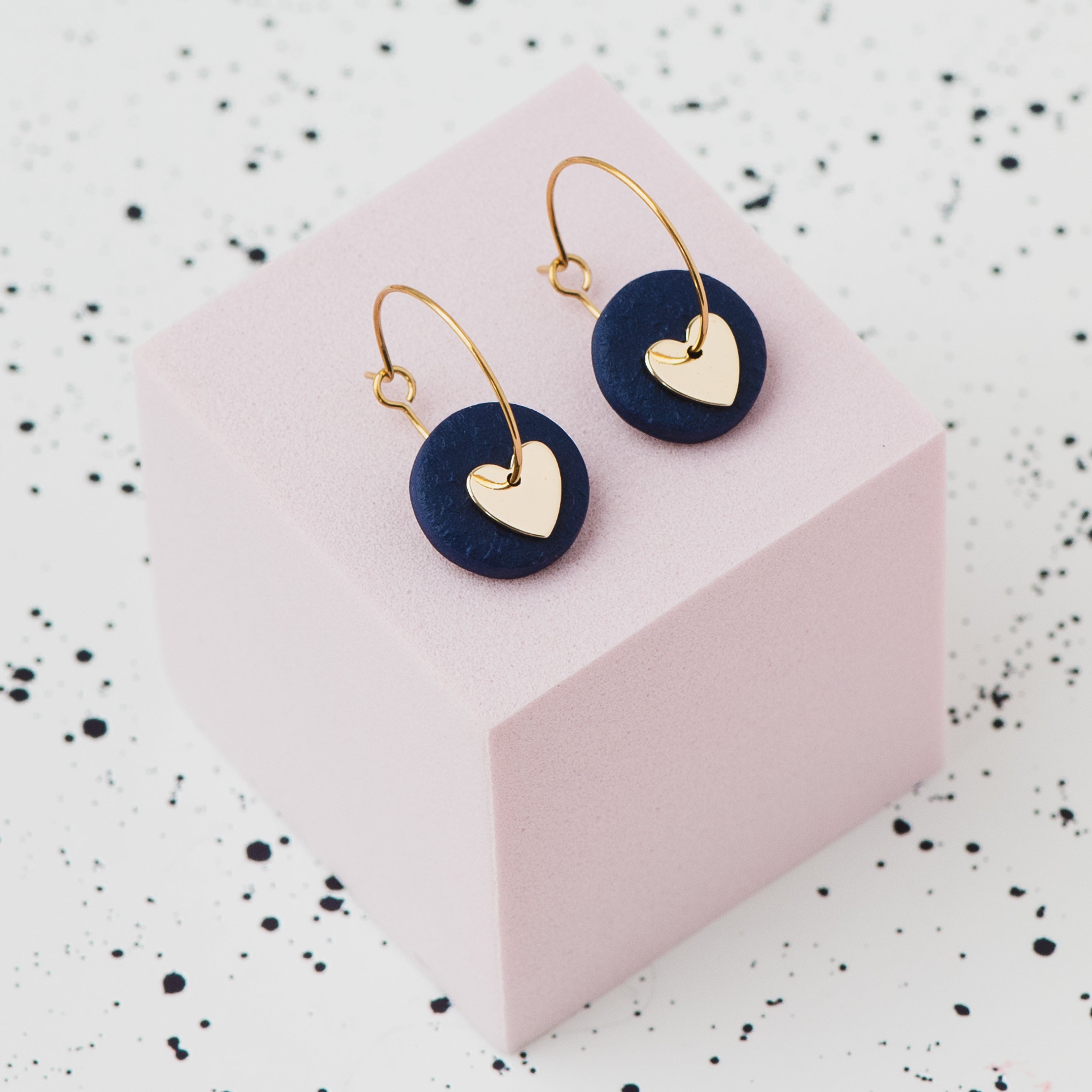 A pair of gold hoop earrings with a navy blue polymer clay disc and a small gold heart, displayed on a light pink cube against a white speckled background.