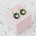 A pair of gold hoop earrings with an olive green polymer clay disc and a small gold heart, displayed on a light pink cube against a white speckled background.