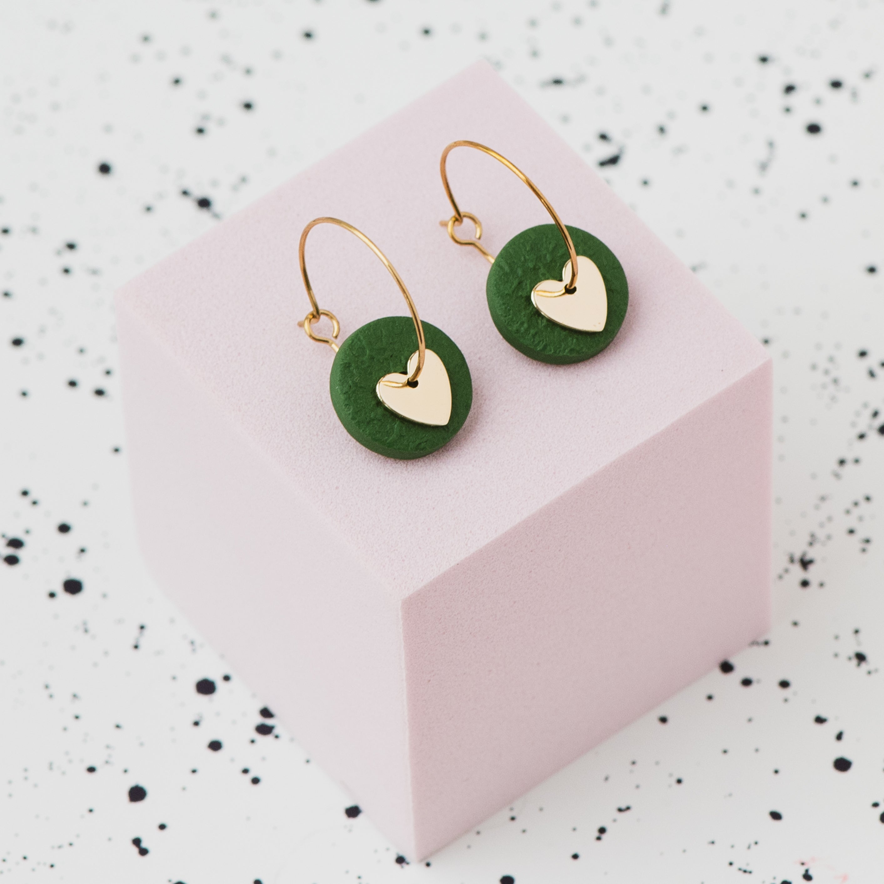 A pair of gold hoop earrings with an olive green polymer clay disc and a small gold heart, displayed on a light pink cube against a white speckled background.