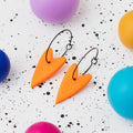 A pair of handmade orange heart-shaped hoop earrings on black hoops, displayed on a white speckled surface surrounded by colourful polymer clay balls.