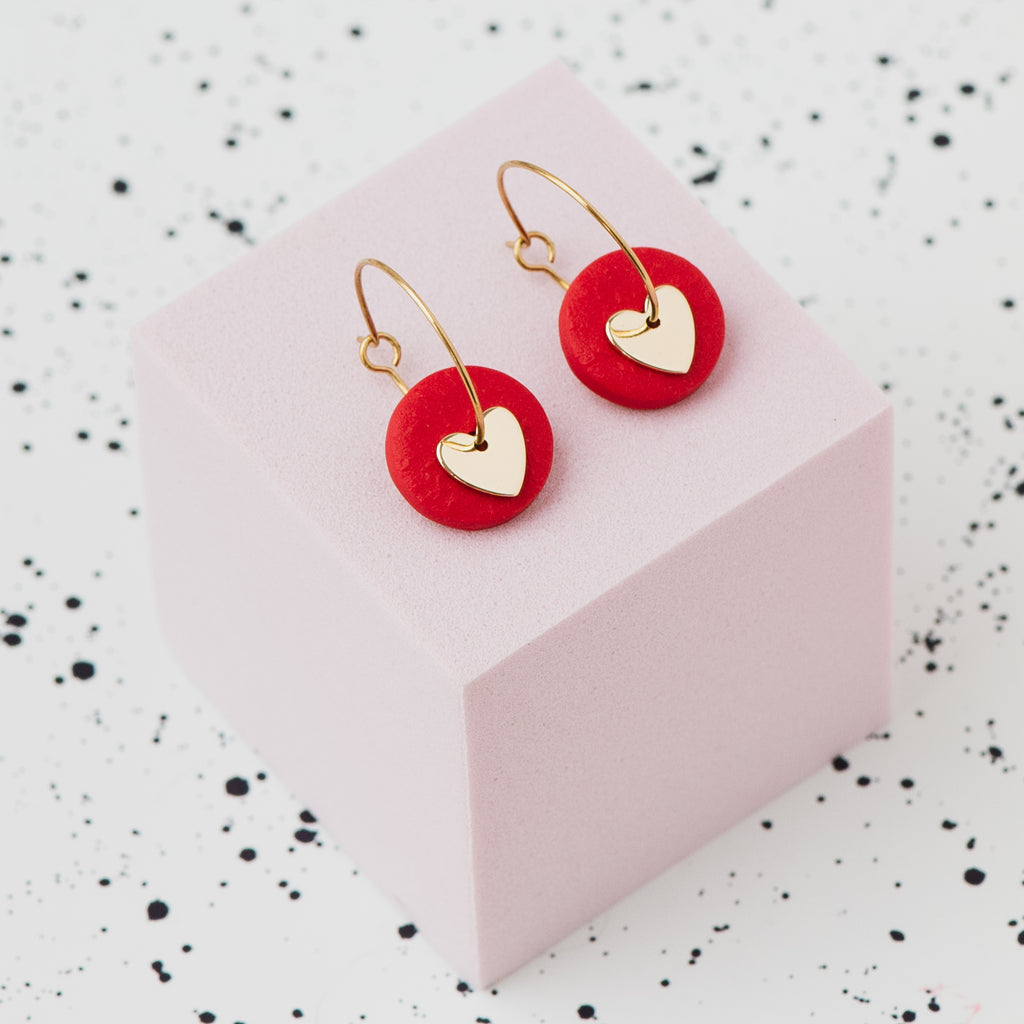 A pair of gold hoop earrings with a red polymer clay disc and a small gold heart, displayed on a light pink cube against a white speckled background.