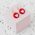 A pair of gold hoop earrings with a red polymer clay disc and a small gold heart, displayed on a light pink cube against a white speckled background.