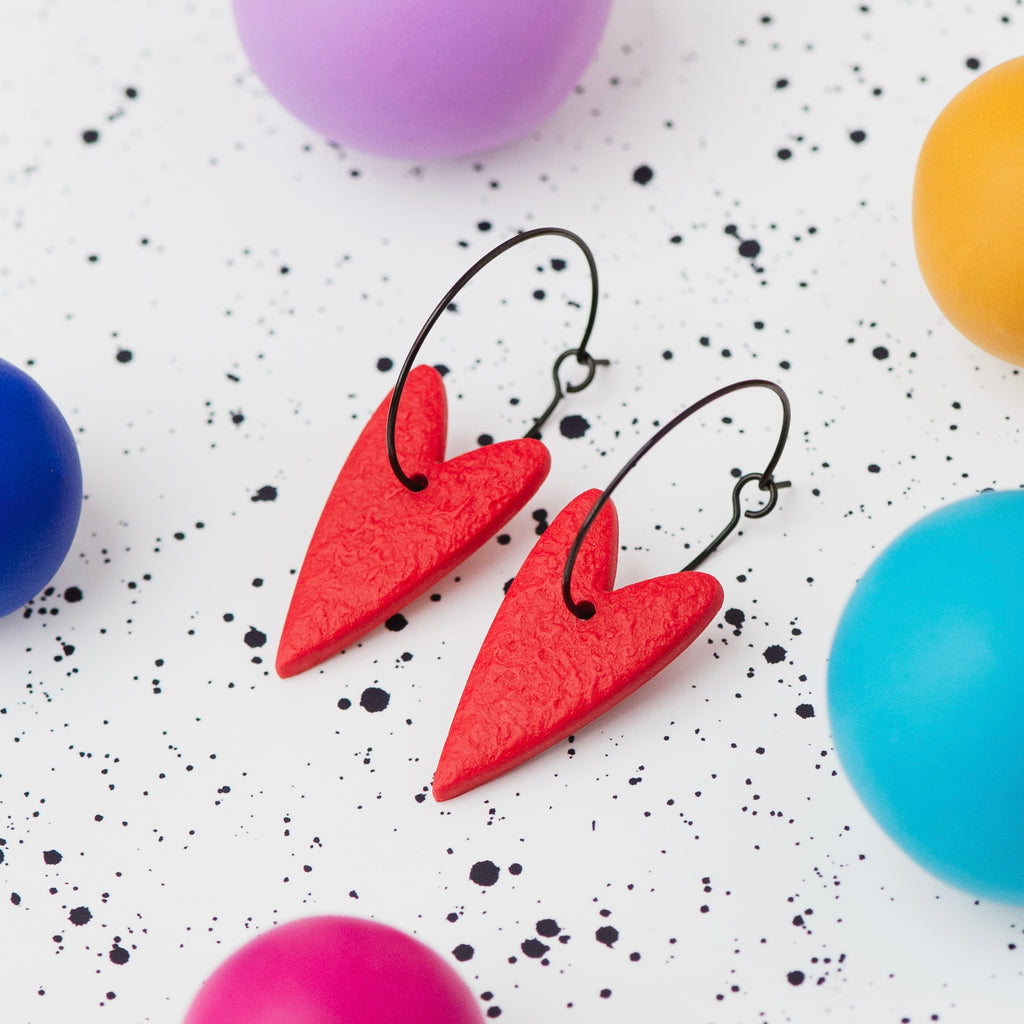 A pair of handmade red heart-shaped hoop earrings on black hoops, displayed on a white speckled surface surrounded by colourful polymer clay balls.